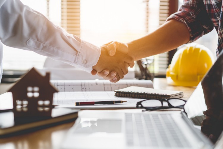 Handshake over desk with model house, glasses and hard hat